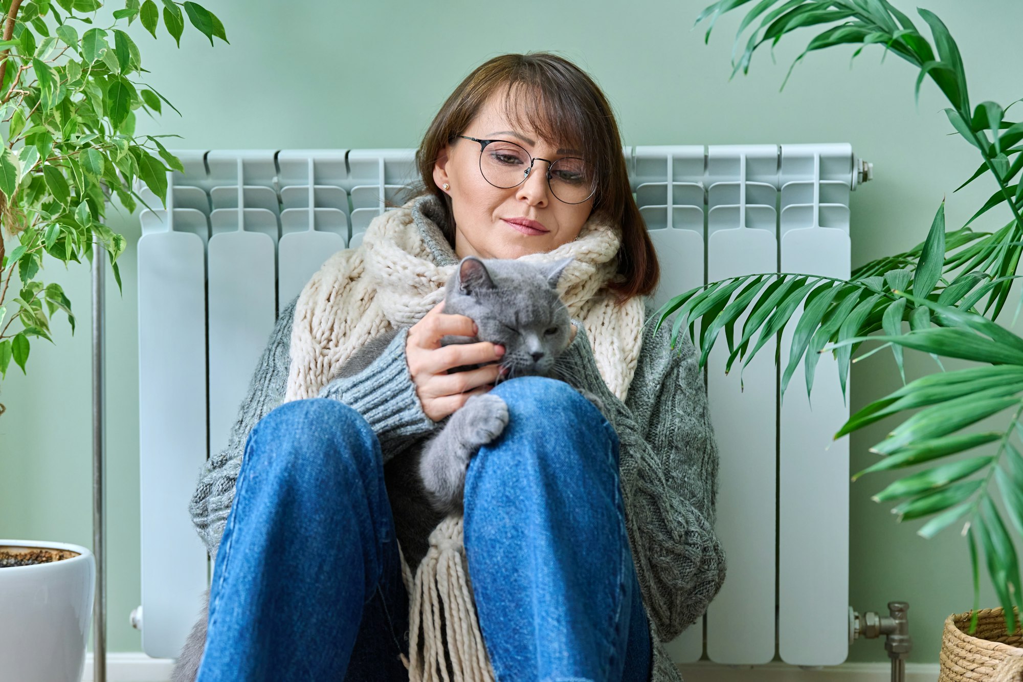 Middle-aged woman in sweater with cat sitting near heating radiator